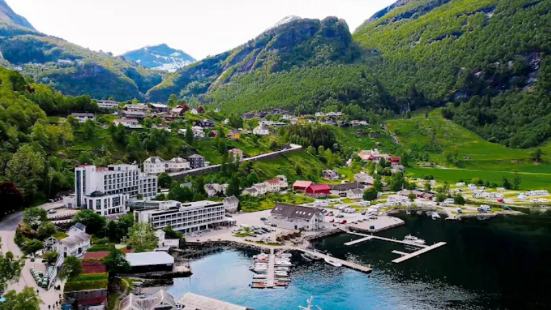 Panoramavy �ver Havila Hotel i Geiranger, Norge, bel�get vid fjorden med sm�b�tshamn, gr�na berg och pittoreska hus i bakgrunden.